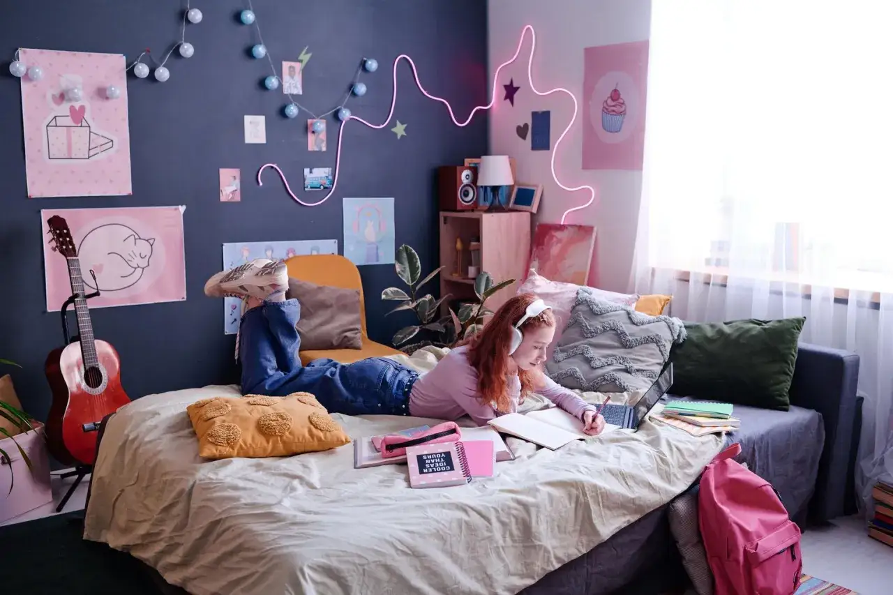 Dziewczyna w słuchawkach na łóżku, otoczona plakatami i lampkami. Pokój młodzieżowy z inspiracjami na kolory ścian: granat, róż, zieleń.