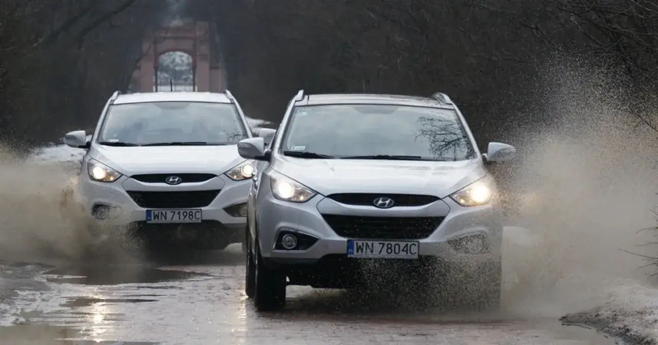Dwa białe Hyundai ix35 pokonują kałużę, rozpryskując wodę.