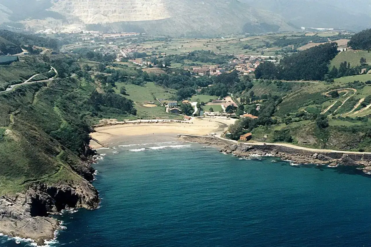 Vista a&eacute;rea de la **playa de Dicido**, con el mar azul turquesa, arena dorada y un pueblo verde en el interior.