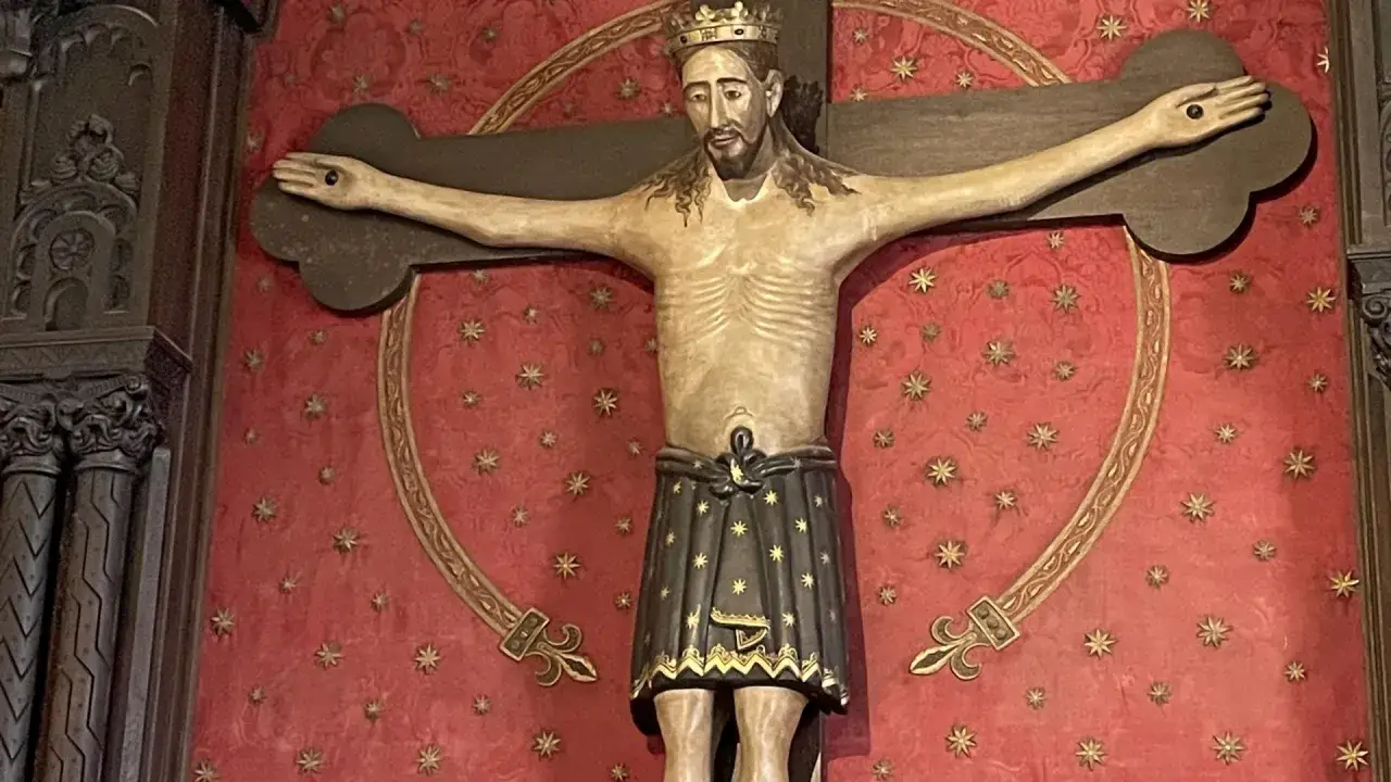 Holzschnitzerei des **Splitters vom Kreuz Jesu** mit Dornenkrone und Lendentuch vor rotem Hintergrund mit goldenen Sternen.