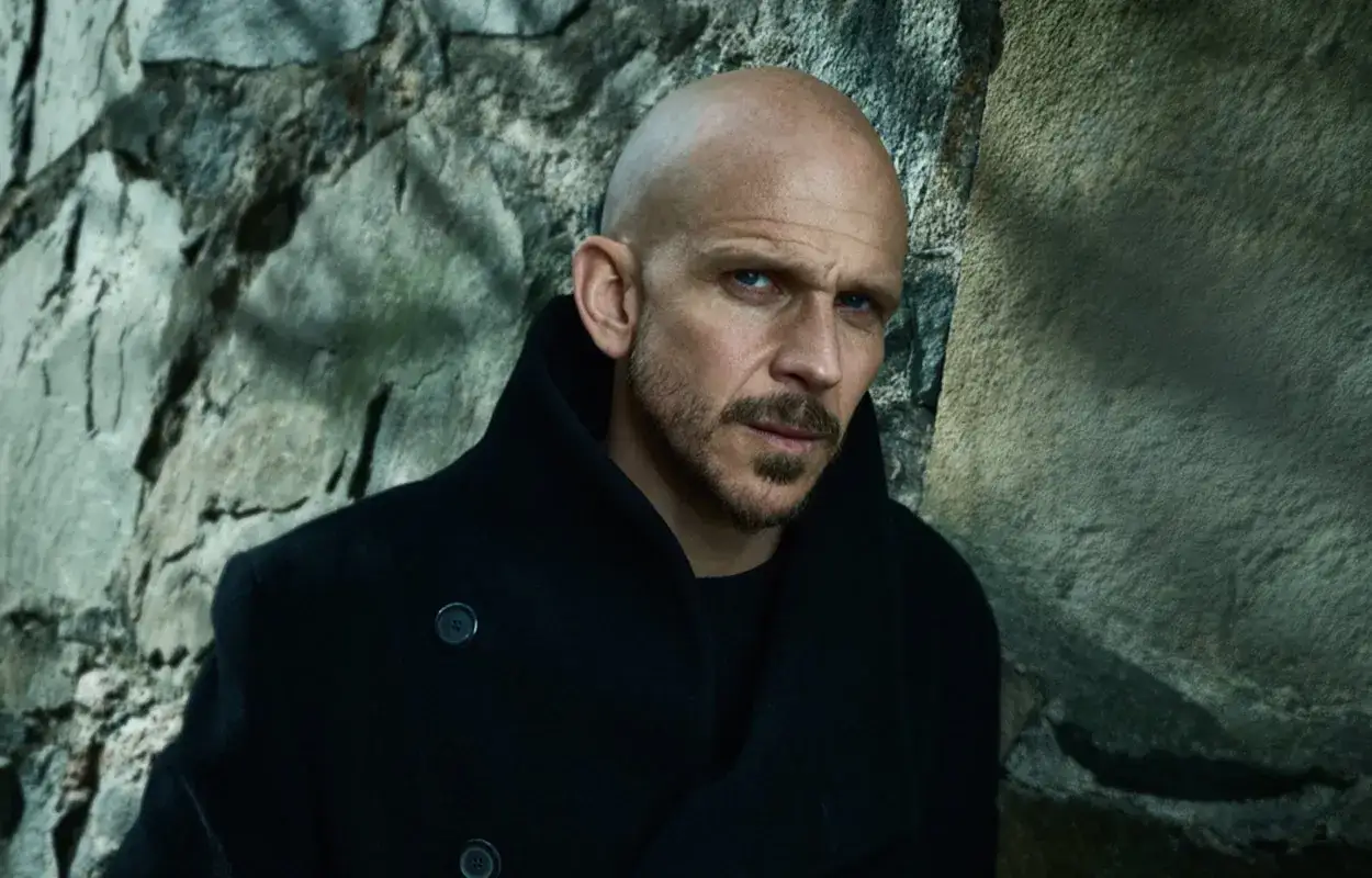 En skallig man med intensiv blick, kl&auml;dd i m&ouml;rk rock, st&aring;r mot en stenv&auml;gg. Han ser ut som en karakt&auml;r fr&aring;n rollistan i Wallander.