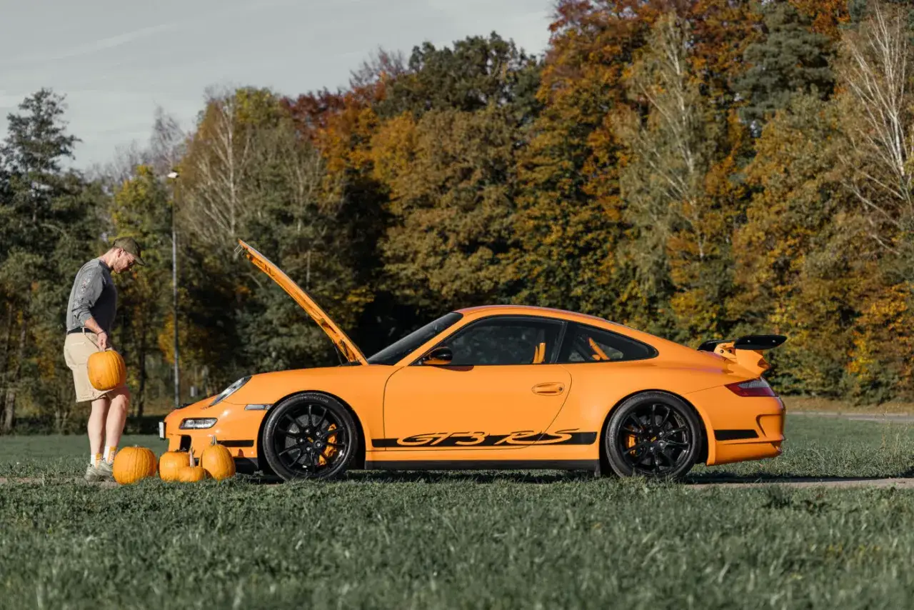 Ein Mann mit Kürbissen steht neben einem leuchtend orangen Porsche 997 GT3 RS im Herbstlaub.