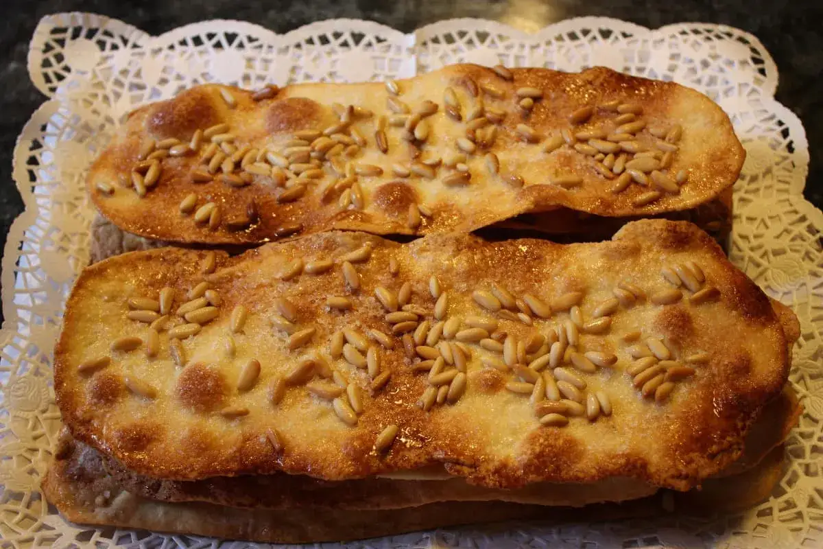 Dos coca de vidre doradas y crujientes, cubiertas de piñones y azúcar, reposan sobre un encaje blanco.