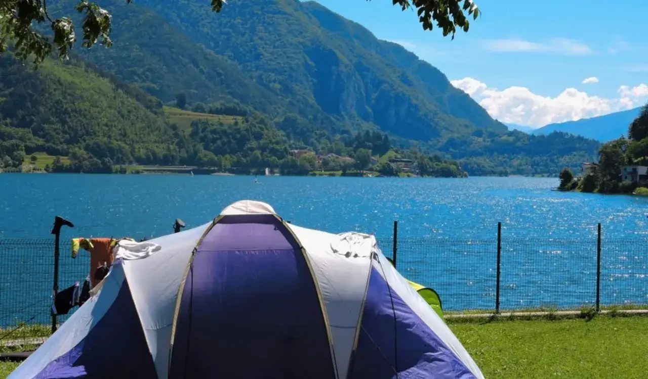 Tenda viola e bianca su prato verde, lago azzurro e montagne sullo sfondo. Il **best Ledro camping** per una vacanza indimenticabile.