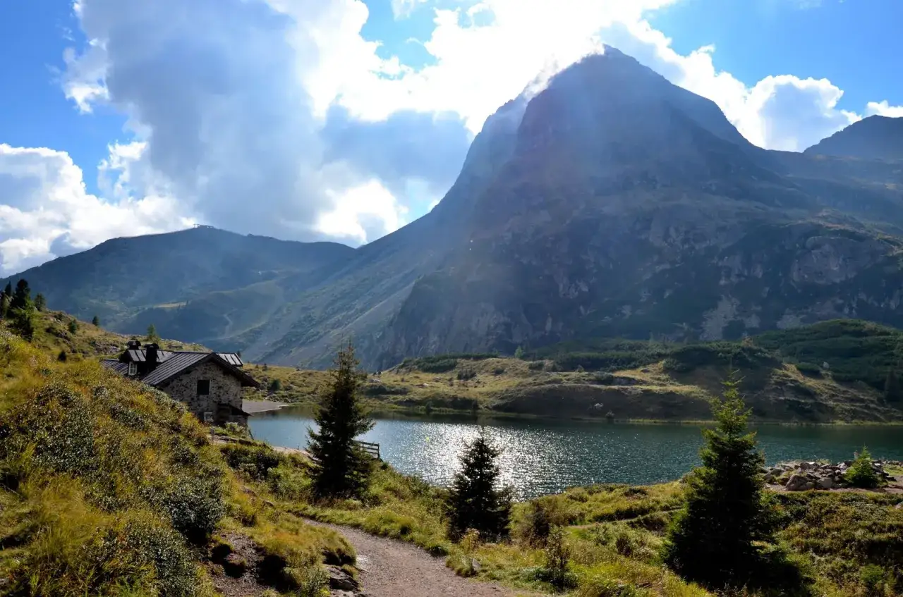 Sentiero per il trekking Lagorai, con baita in pietra e lago alpino sotto imponenti vette.