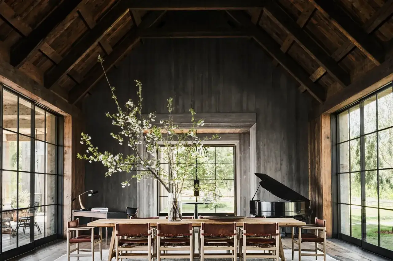 Rustikales Esszimmer mit Holzbalken, Holzw&auml;nden, einem langen Holztisch mit Ledersesseln, einem Fl&uuml;gel und einem bl&uuml;henden Kirschzweig. Ein Hauch von Cottagecore-Charme.