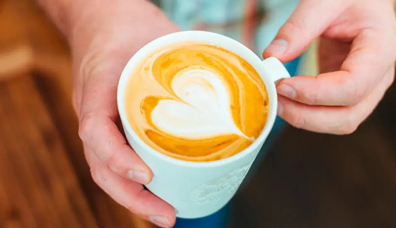 Dłonie trzymają biały kubek z kawą latte, na której widnieje serce z pianki. Idealne latte proporcje dla miłośników kawy.