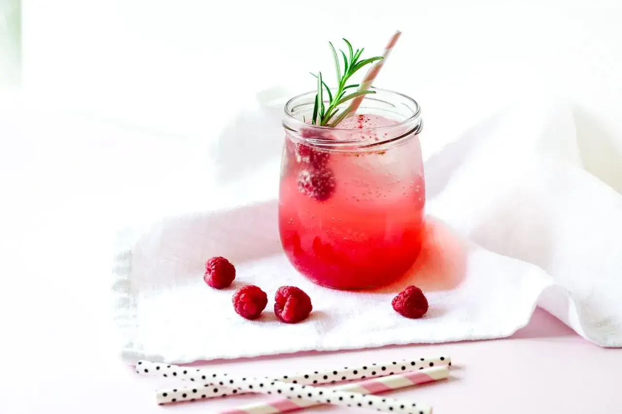 Erfrischender Gin Tonic mit Himbeeren, garniert mit Rosmarin und einem rosa-wei&szlig;en Strohhalm.