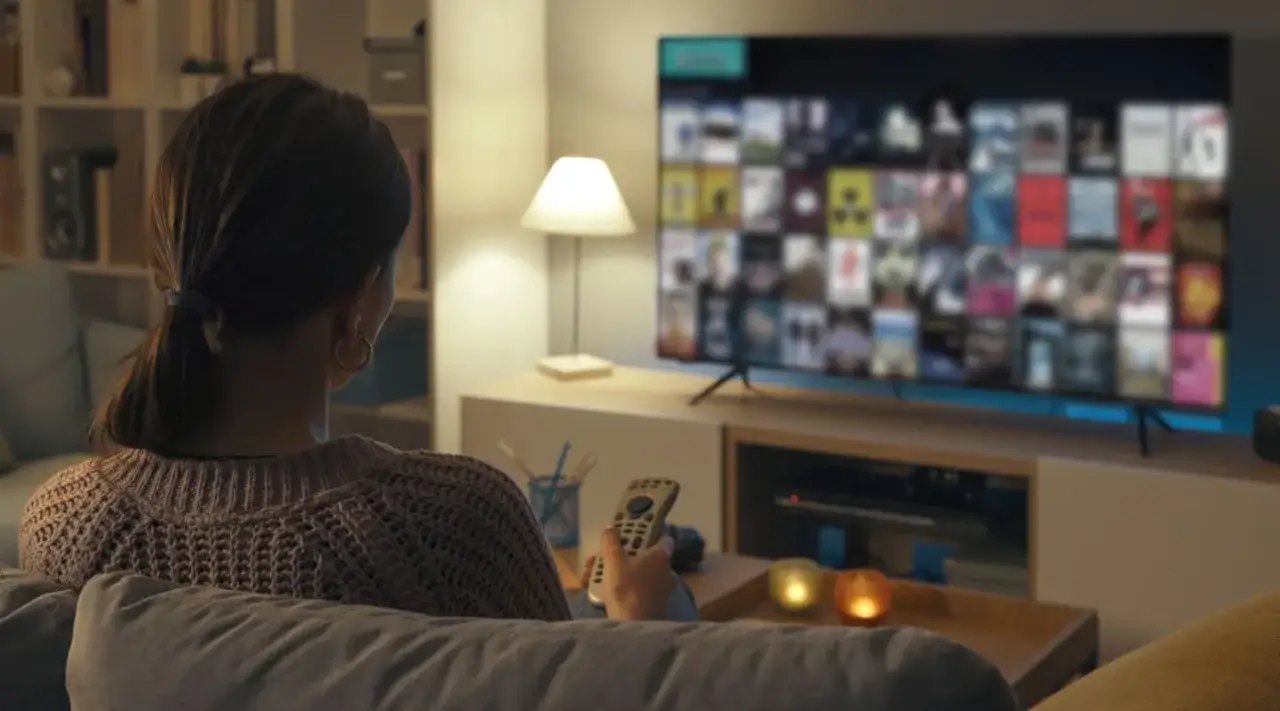 Kobieta ogląda telewizor, zastanawiając się, czy telewizor pobiera dużo prądu. Na ekranie widnieje siatka okładek film&oacute;w.