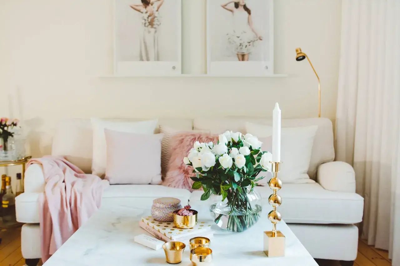 En ljus och elegant soffgrupp med rosa detaljer, som en mjuk pl&auml;d och kuddar. P&aring; soffbordet st&aring;r en vas med vita rosor och ett guldigt ljusstake, en inredningsstil som p&aring;minner om Lovisa Barkman.