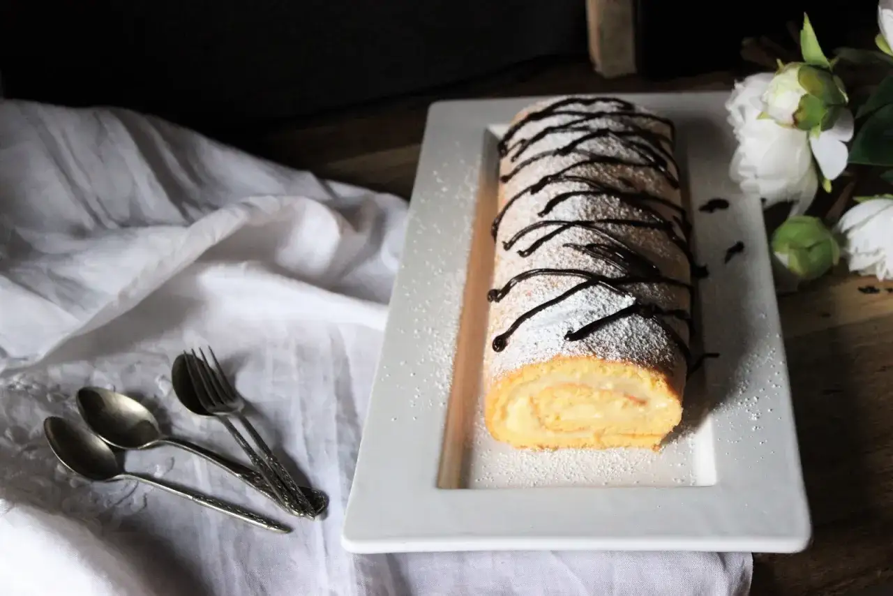 Un delicioso brazo de gitano cubierto de azúcar glas y chocolate, listo para ser disfrutado con cucharas y tenedor.
