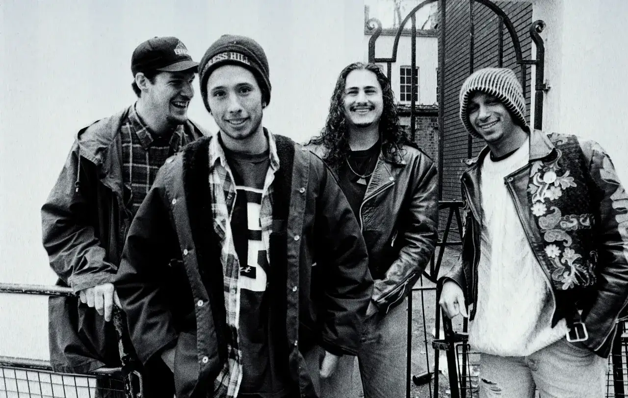 Członkowie Rage Against the Machine pozują do zdjęcia. Wszyscy uśmiechają się, ubrani w charakterystyczne dla lat 90. kurtki i czapki.