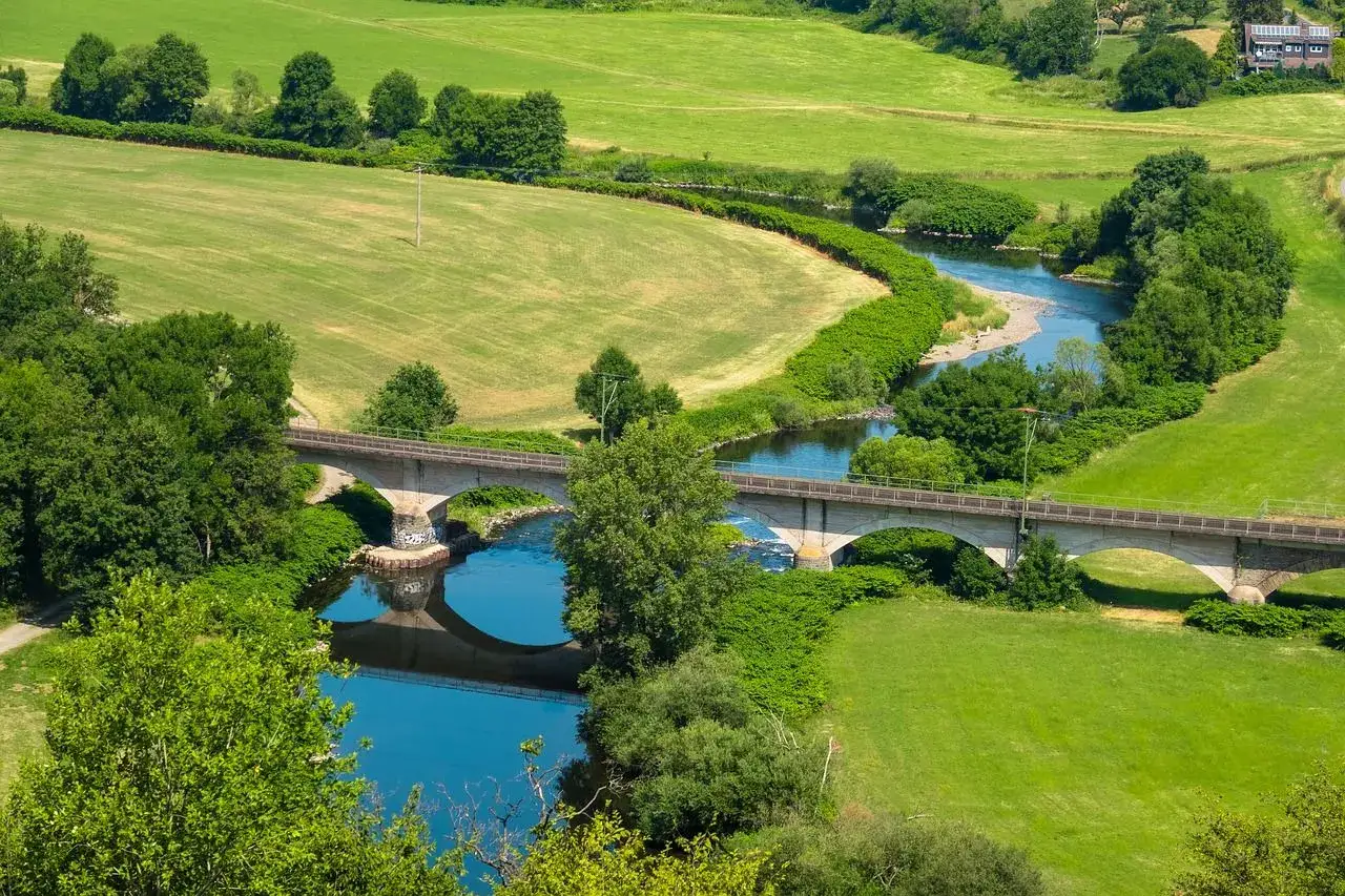 Gr&uuml;ne Wiesen und eine geschwungene Sieg laden zum Wandern ein. Eine Eisenbahnbr&uuml;cke &uuml;berspannt den Fluss, der sich durch die Landschaft schl&auml;ngelt.