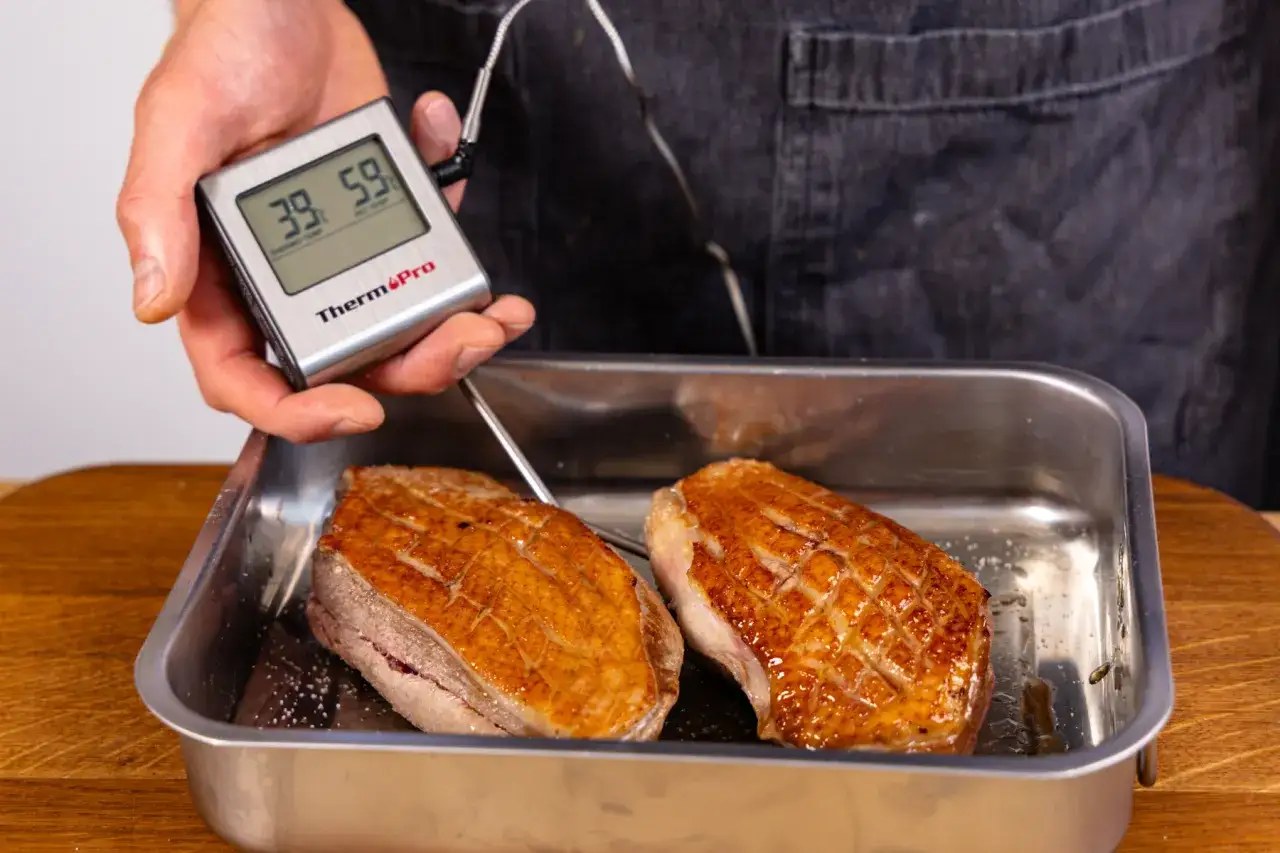 Perfekte kerntemperatur entenbrust: Ein Thermometer misst 39&deg;C und 59&deg;C bei zwei gebratenen Entenbr&uuml;sten in einer Metallschale.
