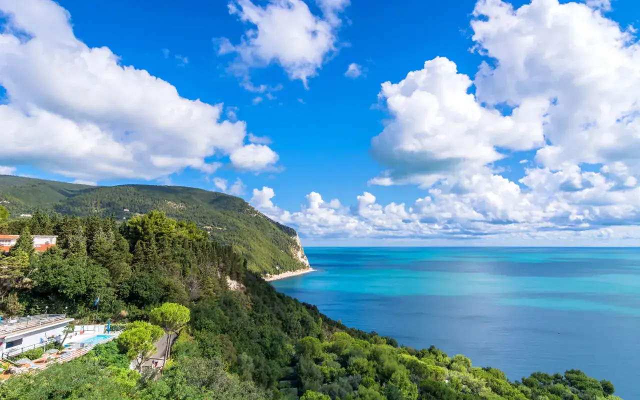 Splendido **paesaggio** delle **Marche**: costa verdeggiante, mare azzurro e cielo con nuvole bianche.