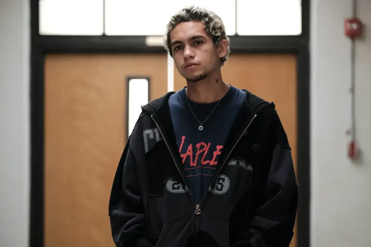 Fezco, personnage de la saison 2 d'Euphoria, pose dans un couloir, portant une veste zippée noire sur un t-shirt bleu.