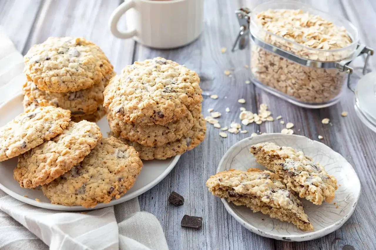 Des cookies flocons d'avoine, certains empil&eacute;s sur une assiette blanche, d'autres coup&eacute;s en deux pour r&eacute;v&eacute;ler leur c&oelig;ur chocolat&eacute;. Un pot de flocons d'avoine est visible en arri&egrave;re-plan.