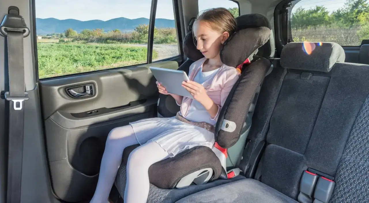 Niña en alzador coche, cumpliendo la normativa de seguridad vial, entretenida con una tablet.