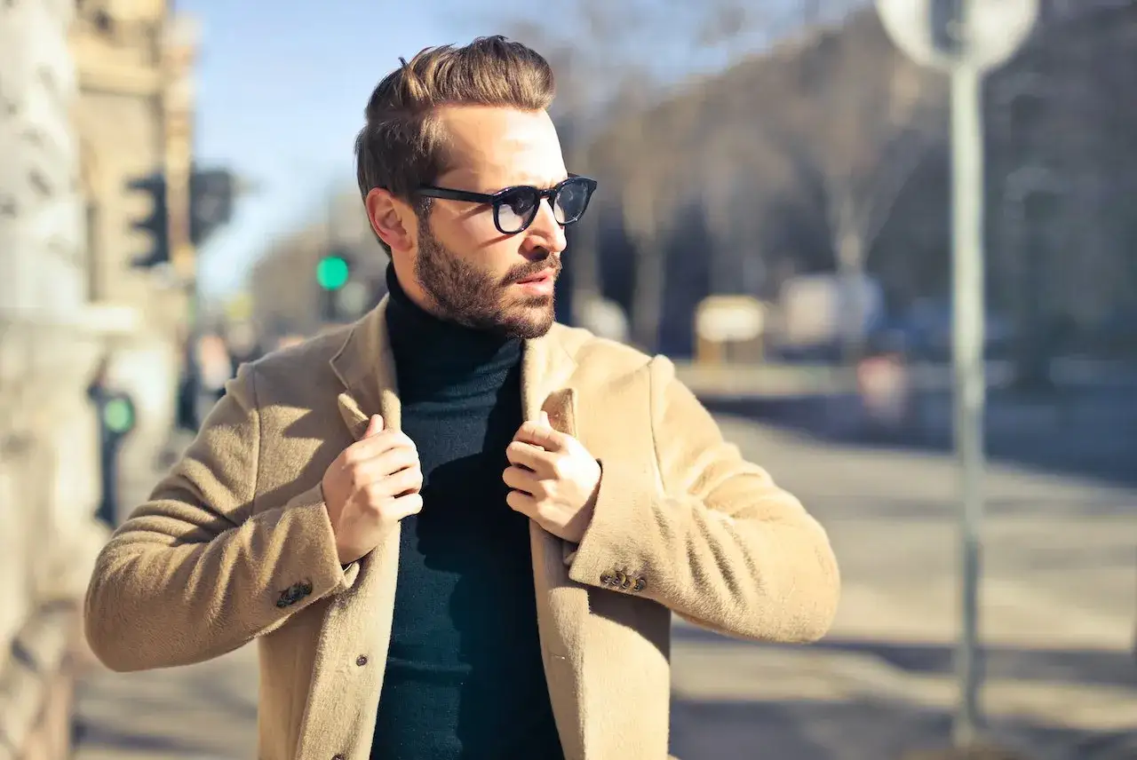 Ein Mann mit Bart und Sonnenbrille, der sich stilvoll kleidet, trägt einen beigen Mantel über einem schwarzen Rollkragenpullover.