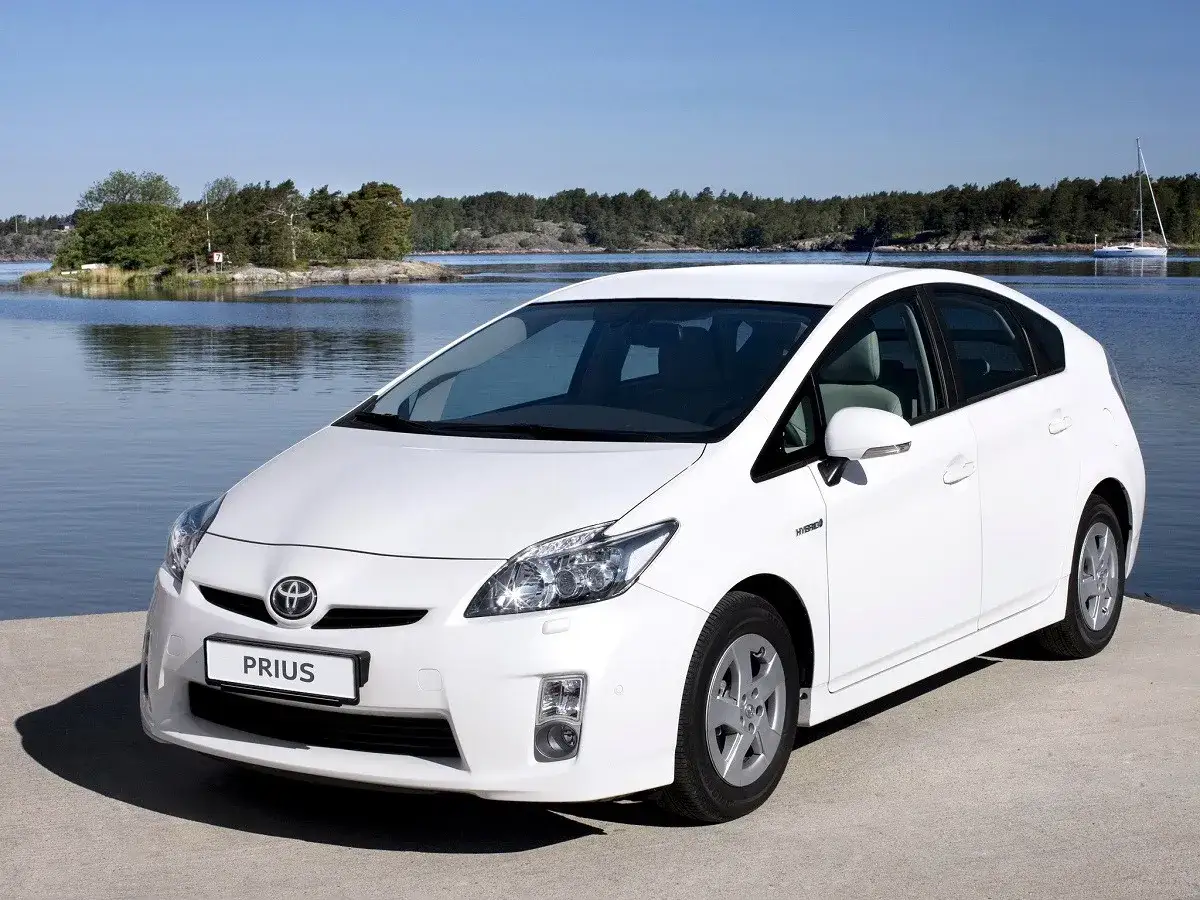 Biały Toyota Prius 3 zaparkowany nad wodą, z wyspą i żaglówką w tle.