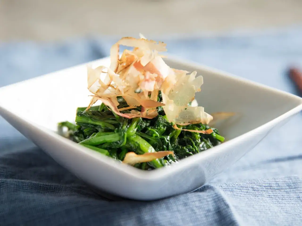 A white bowl holds wilted spinach topped with delicate bonito flakes, hinting at a savory ankake sauce.
