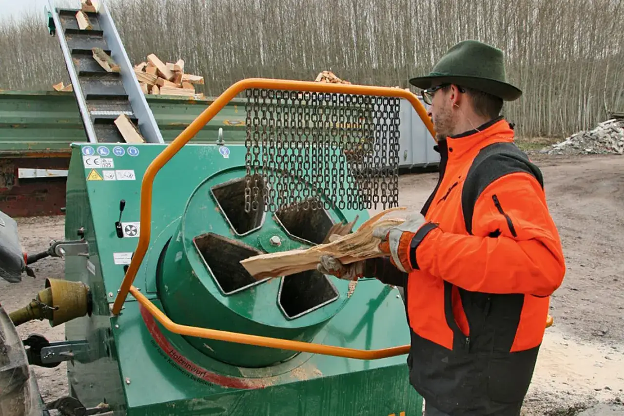 Mann mit orangefarbener Jacke und Hut füttert Holz in einen Holzhäcksler mit einem 4m Förderband für Holz.