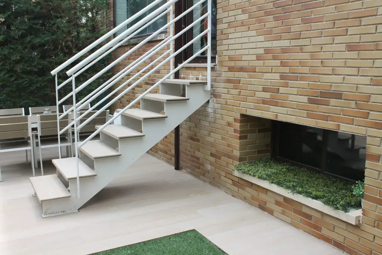 Escalera abierta al exterior con barandillas blancas, junto a una pared de ladrillos y un jard&iacute;n exterior.