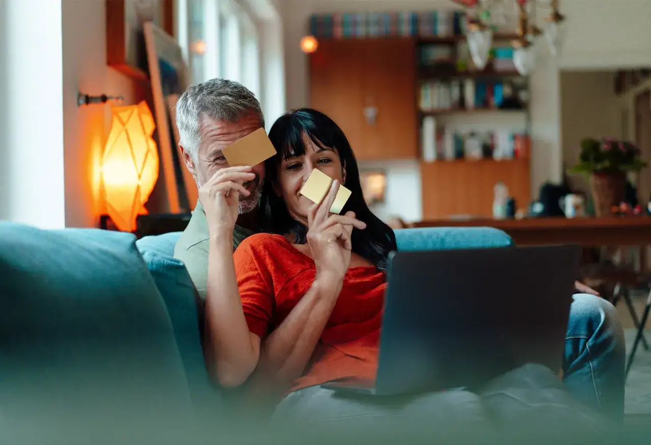 Pareja sonriente con tarjetas doradas, planeando sus finanzas en su cuenta conjunta mientras miran una laptop.