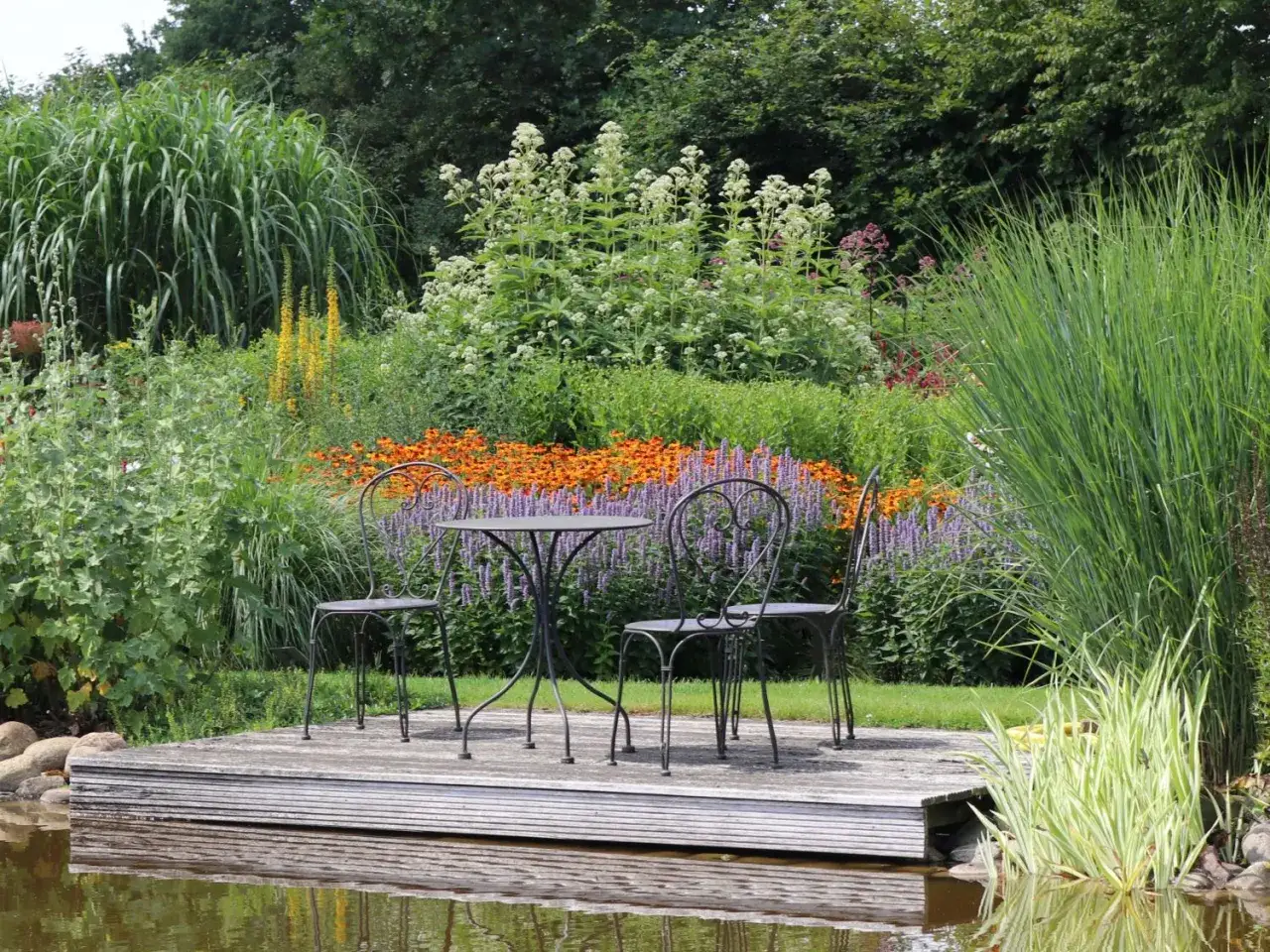 Ein idyllischer **Wasserdost Standort** mit einem kleinen Tisch und zwei Stühlen auf einer Holzplattform am Teich. Üppige Blumen und Gräser umgeben die Szenerie.