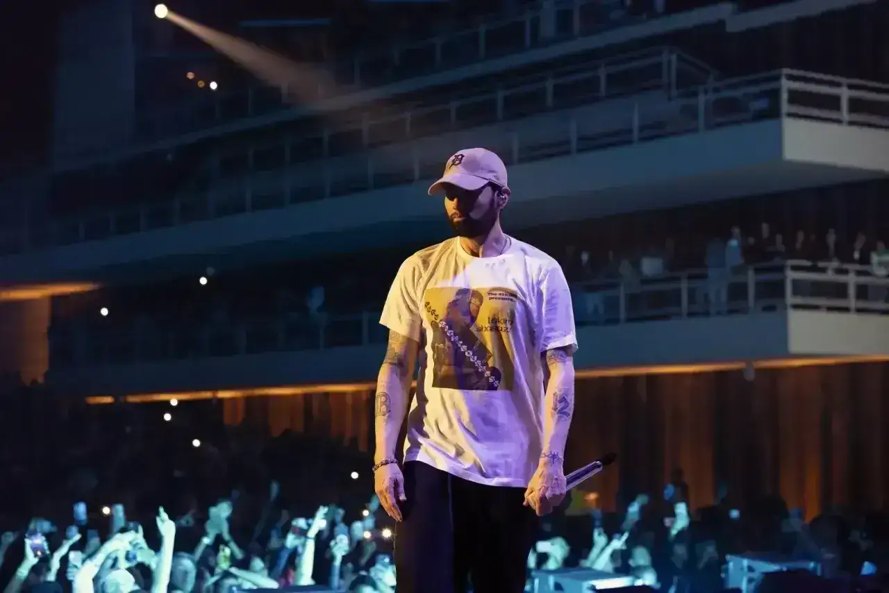 Eminem, jako muzyk, stoi na scenie w czapce i koszulce z nadrukiem, trzymając mikrofon. Tłum widzów z telefonami wznosi ręce.
