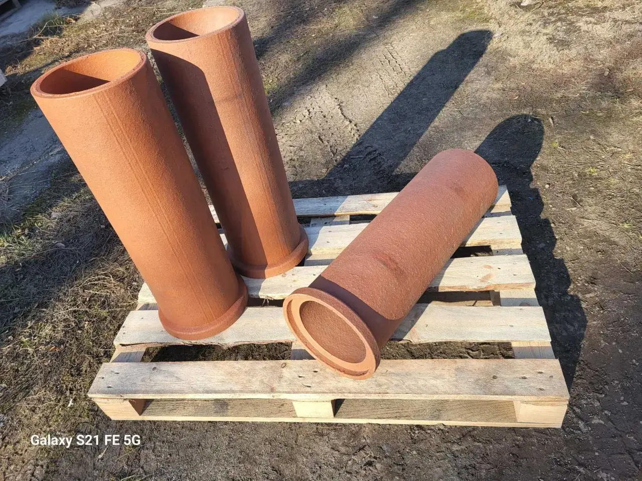 Trzy rdzawobrązowe, ceramiczne rury, stanowiące kamionkowy materiał budowlany, leżą na drewnianej palecie.