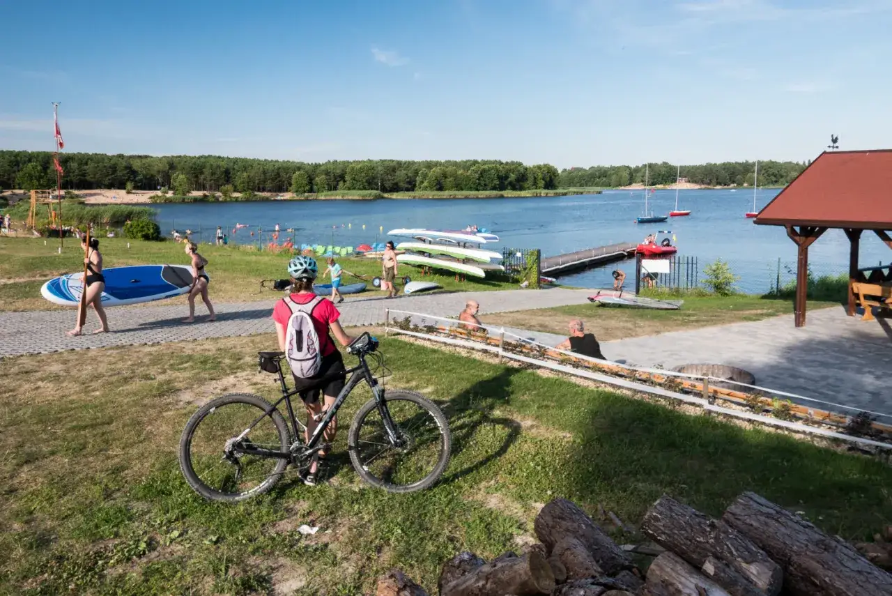 Letni dzień nad zalewem Cedzyna. Ludzie spacerują, niosą deski SUP, rower czeka na właścicielkę. W tle jezioro, łódki i las.