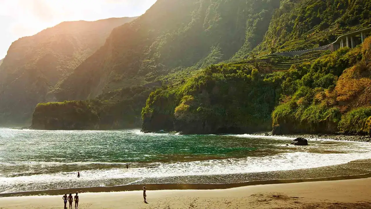 Ein malerischer madeira schwarzer strand mit sanften Wellen und &uuml;ppig gr&uuml;nen Bergen im Hintergrund. Ein paar Menschen genie&szlig;en die Sonne.