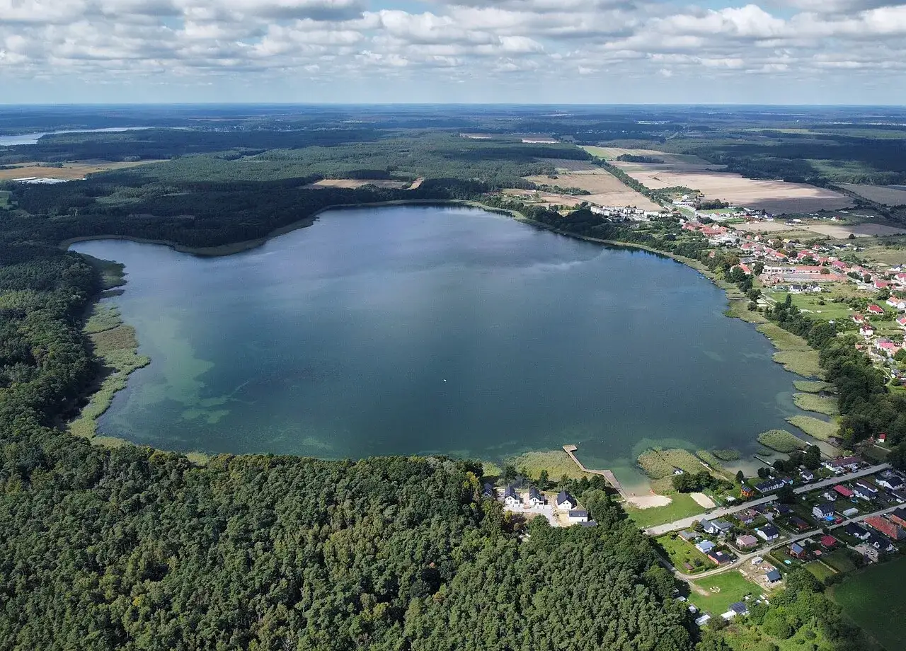 Malownicze jezioro Wilkowskie otoczone lasami i zabudowaniami. Woda jest spokojna, a na brzegu widać domy i plażę.