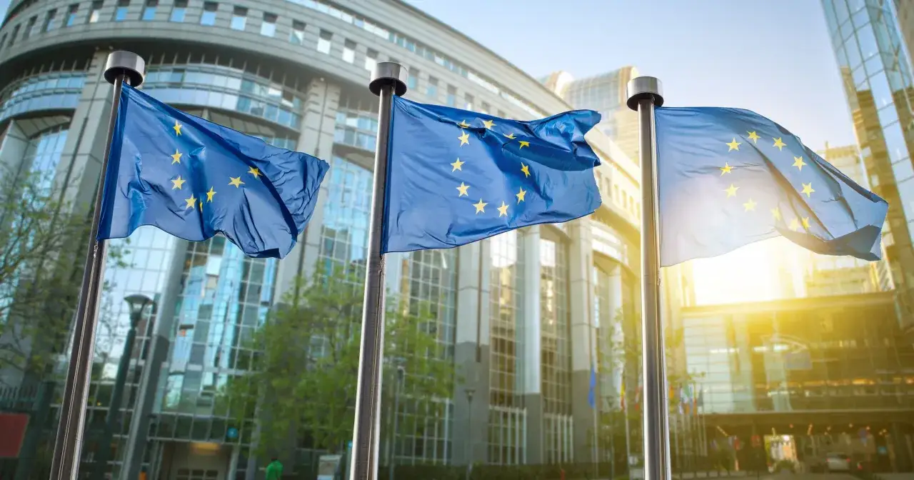 Flagi Unii Europejskiej powiewają przed nowoczesnym budynkiem. Symbolizują wspólne wartości i aksjologię Europy.