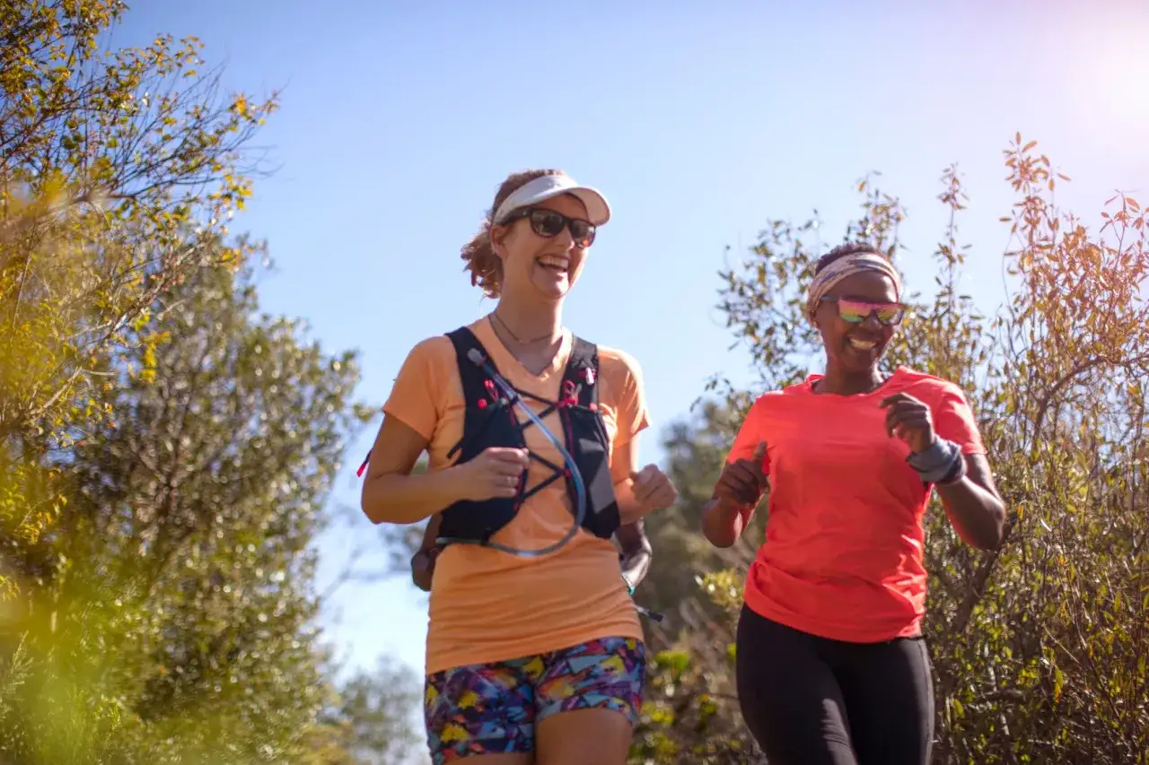 Deux femmes souriantes s'entra&icirc;nent en plein air, profitant de la nature. Leur **m&eacute;thode norv&eacute;gienne course** les pousse &agrave; se d&eacute;passer.