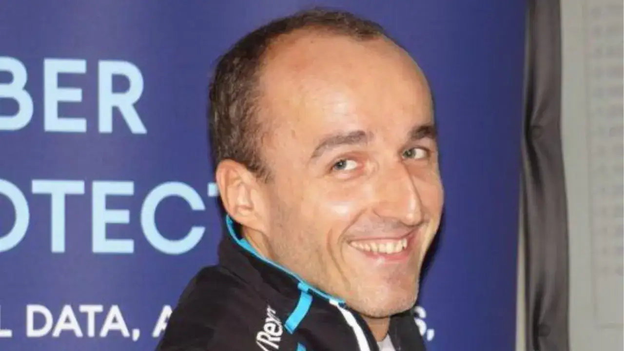 Uśmiechnięty Robert Kubica, który niedawno został ojcem, pozuje do zdjęcia na tle niebieskiego banera.