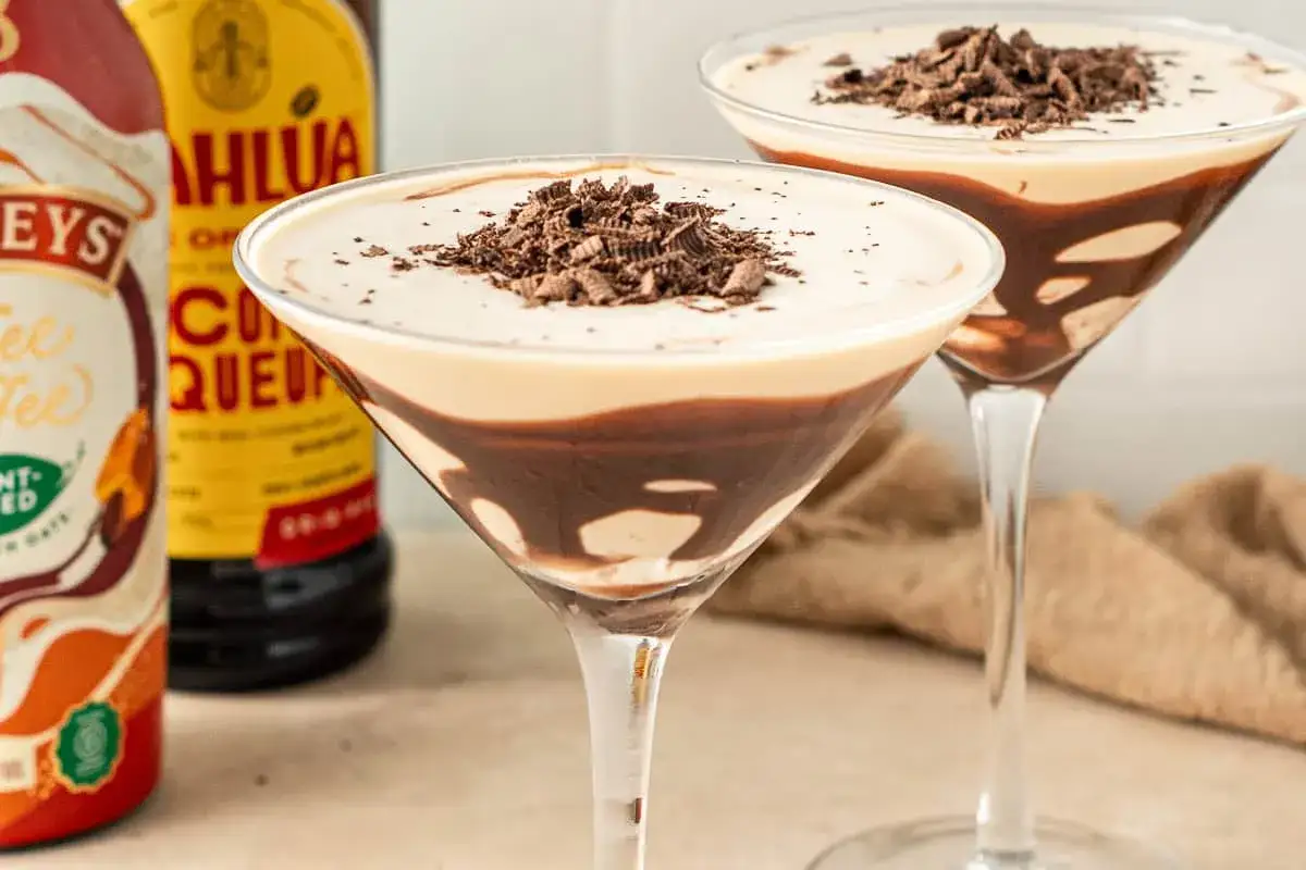 Två martiniglas med en krämig mudslide drink, toppad med chokladspån. Bredvid syns flaskor med Kahlúa och Baileys.