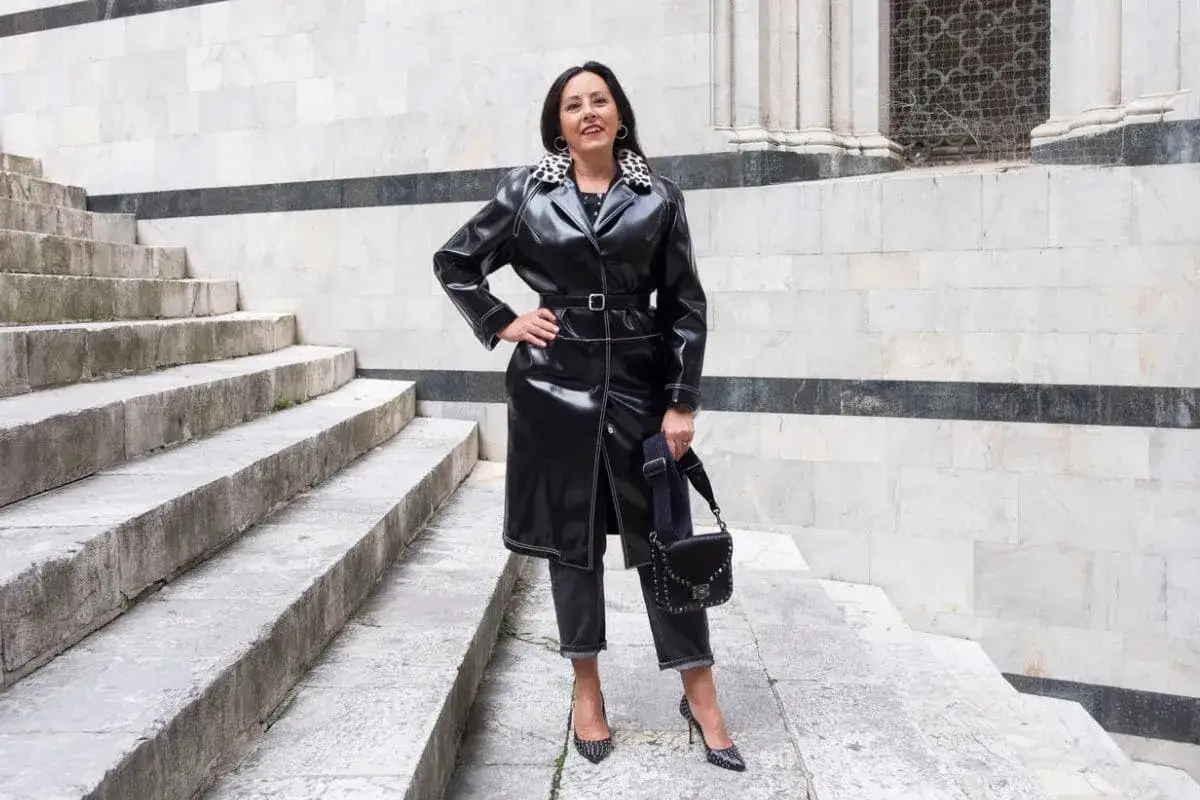 Donna con un audace outfit rock: trench in ecopelle nera, jeans scuri, tacchi borchiati e borsa a tracolla.