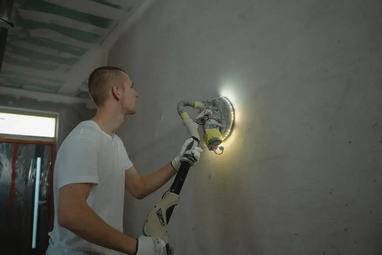 Hombre con guantes blancos lijando una pared con una máquina. La herramienta tiene luces LED que iluminan la superficie.