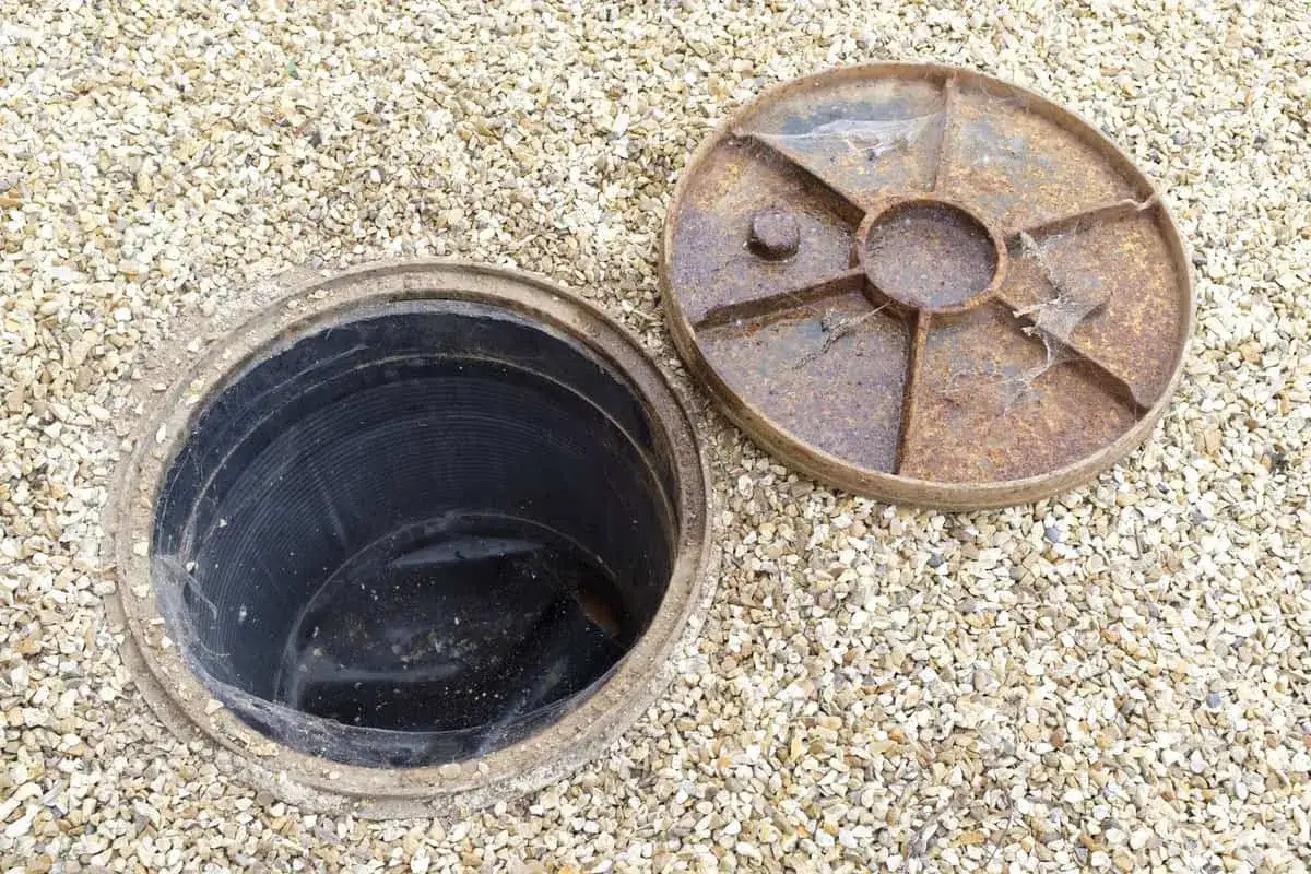 Inspection d'une canalisation bouch&eacute;e sous la maison. Le couvercle rouill&eacute; est retir&eacute;, r&eacute;v&eacute;lant le conduit noir.