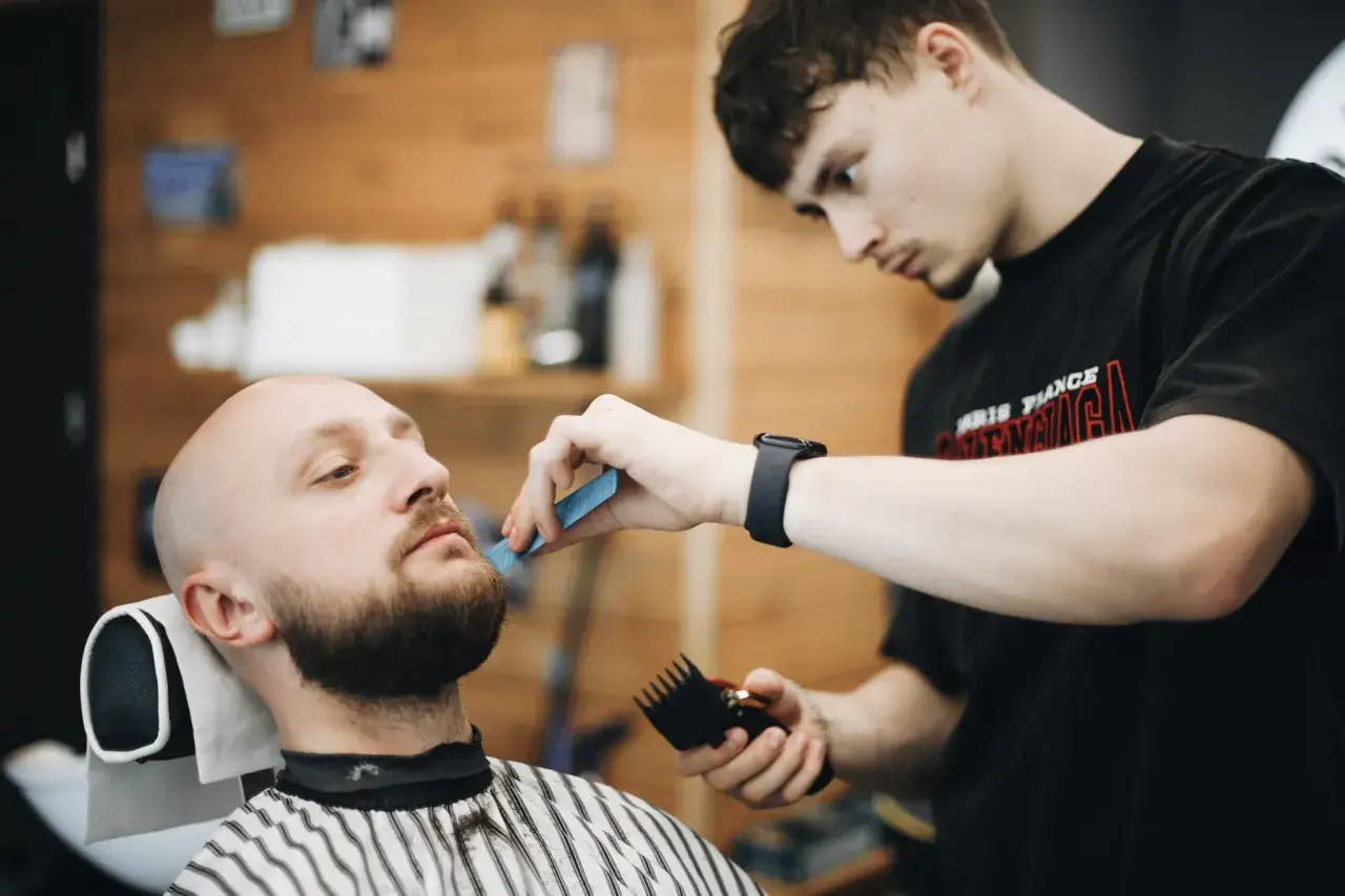 Młody barber z grzebieniem i maszynką precyzyjnie przycina brodę klienta w salonie.