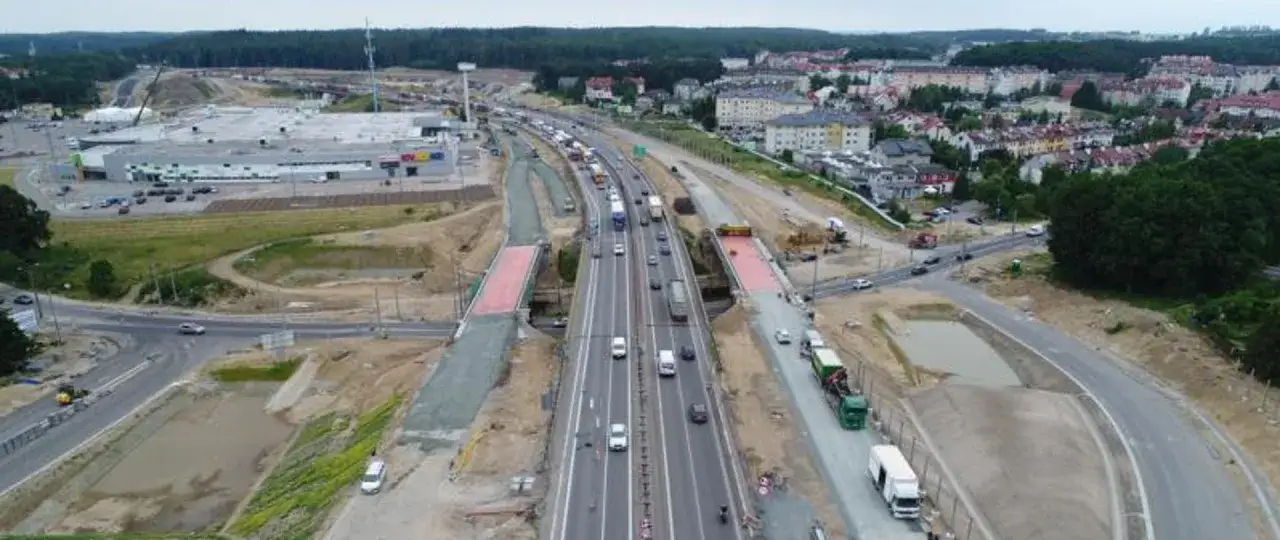Budowa trasy kaszubskiej widać ruchliwy odcinek drogi z samochodami i ciężarówkami, a także nowe wiadukty i tereny budowy.