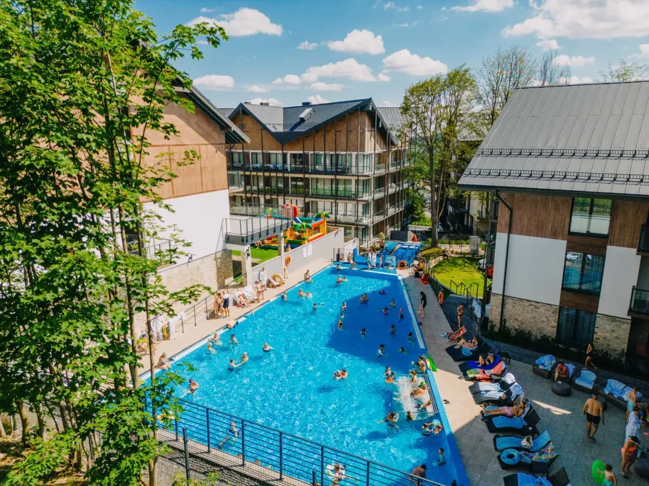 Hotel z podgrzewanym basenem zewnętrznym, gdzie rodziny cieszą się słońcem i wodą. Dzieci bawią się na dmuchanych zjeżdżalniach, a dorośli relaksują się na leżakach.