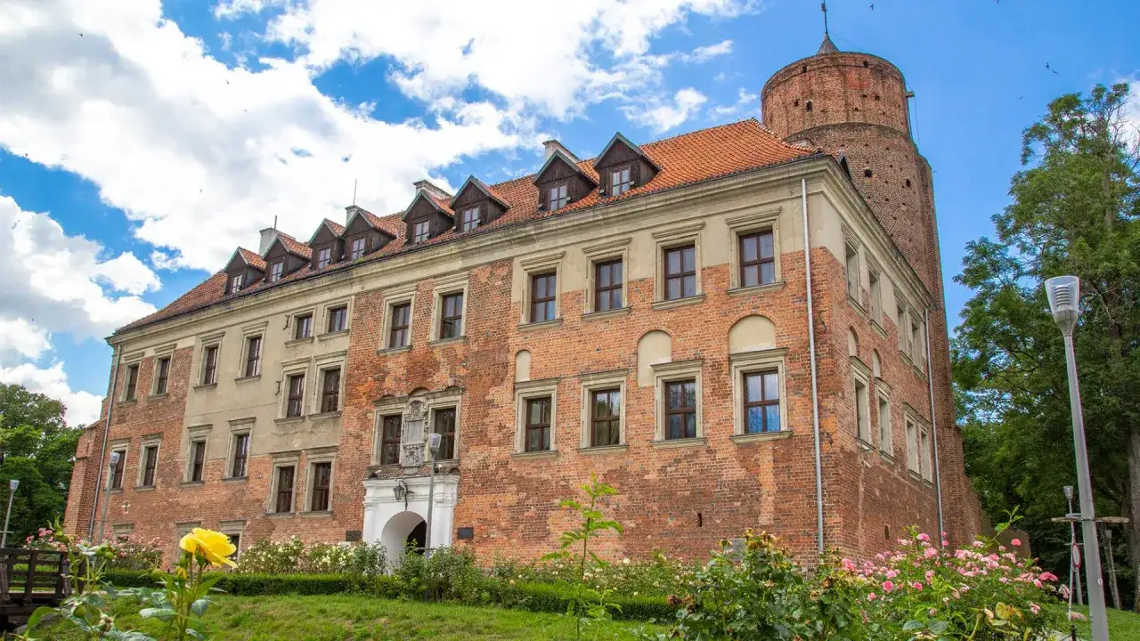 Zamek w Uniejowie i okolice atrakcje. Ceglana budowla z wieżą, otoczona zielenią i kwitnącymi różami, zaprasza do odkrywania historii.