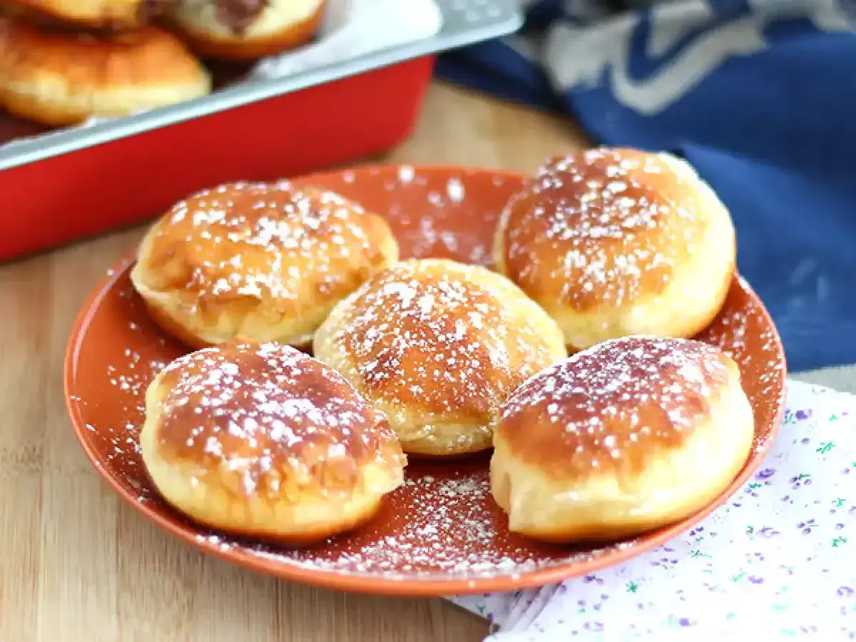 Cinq beignets au chocolat saupoudr&eacute;s de sucre glace sur une assiette orange. Un beignet au chocolat est visible dans un plat rouge en arri&egrave;re-plan.