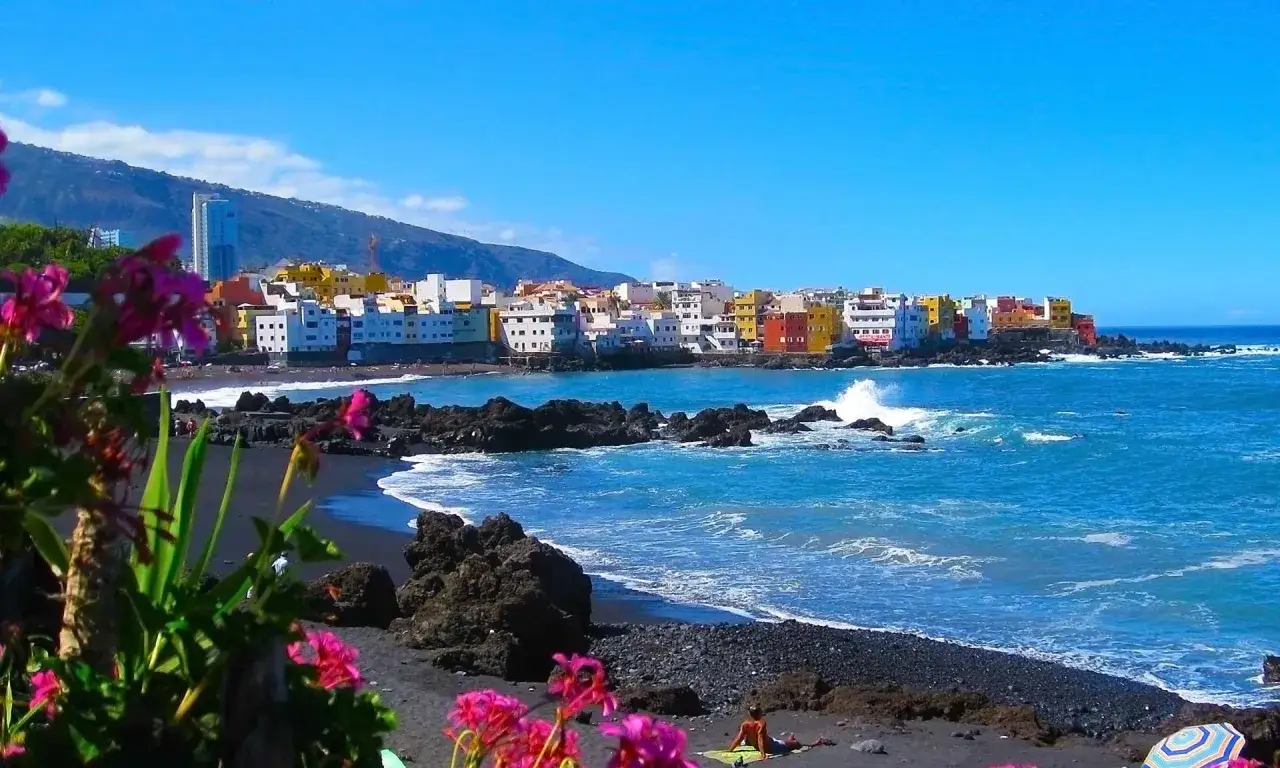 Pueblo costero de casas coloridas, playa de arena negra y flores rosas. Un lugar ideal para saber donde viajar en julio barato.