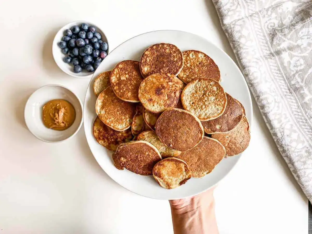 Ein Teller voller kleiner, goldbrauner Baby Pancakes, daneben eine Schüssel mit Blaubeeren und Erdnussbutter. Perfekt für einen süßen Start in den Tag!