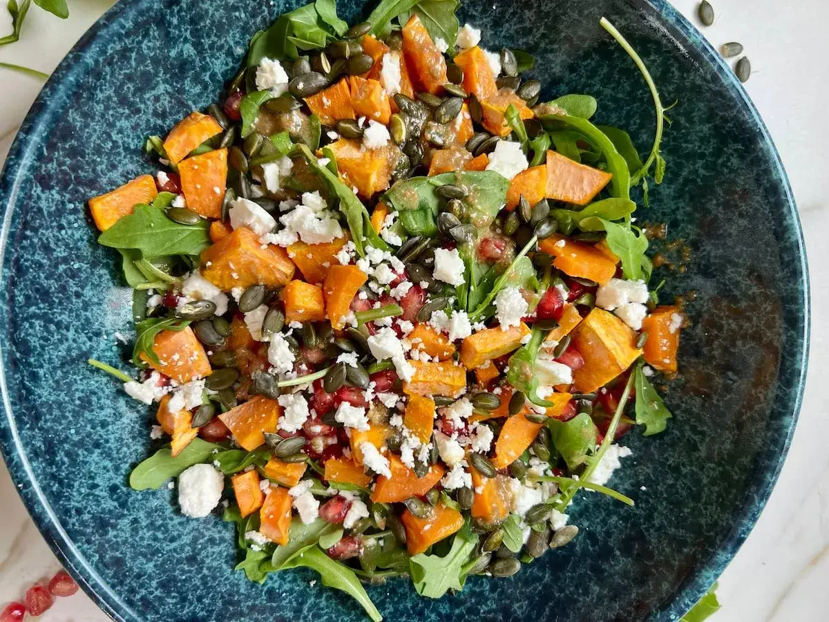 Ensalada vibrante con boniato asado, queso feta, granada y semillas de calabaza sobre rúcula. Perfecta para tus recetas con boniato.