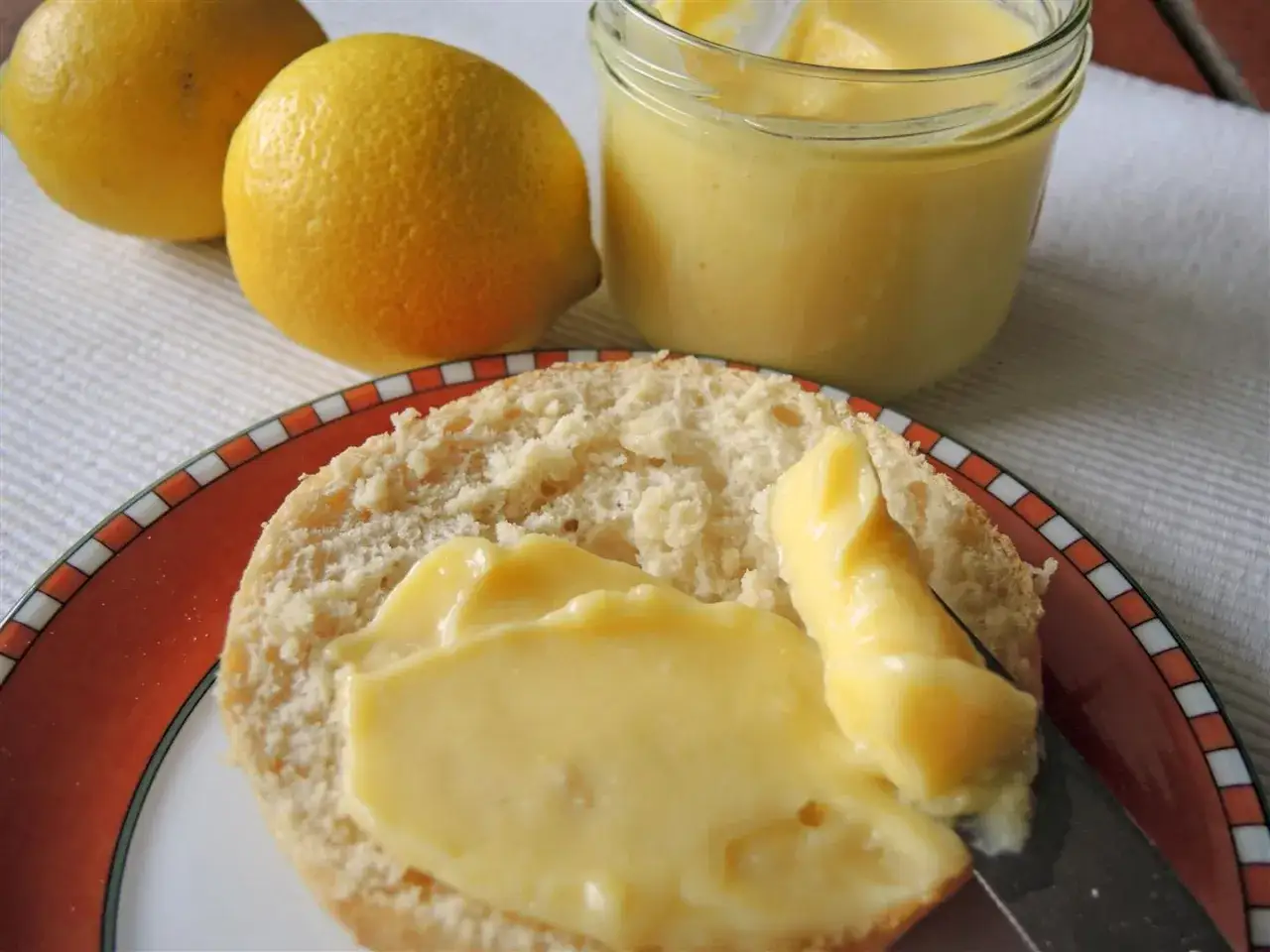 Ein Brötchen wird mit cremigem Lemon Curd bestrichen. Daneben liegen frische Zitronen und ein Glas mit dem süß-säuerlichen Aufstrich.